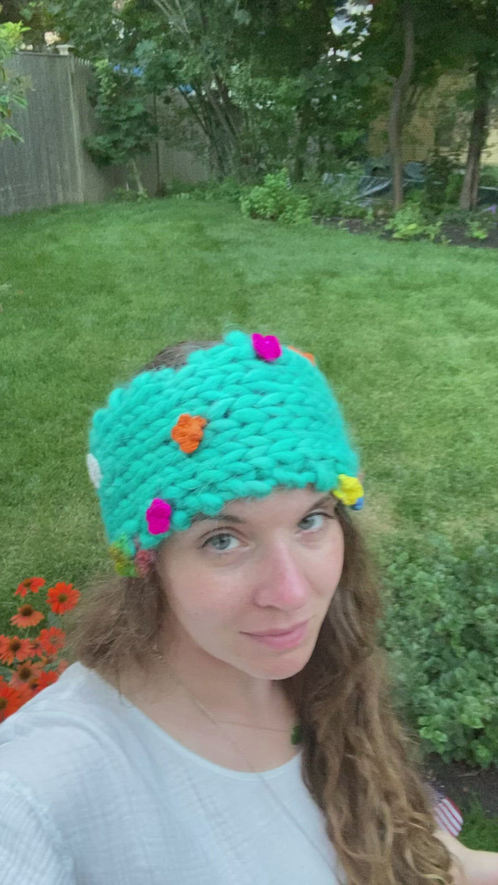Video of founder Nicole wearing flower crown wool headband outside with grass and trees in the background. Headband is knit, with many bright colored flowers interspersed.