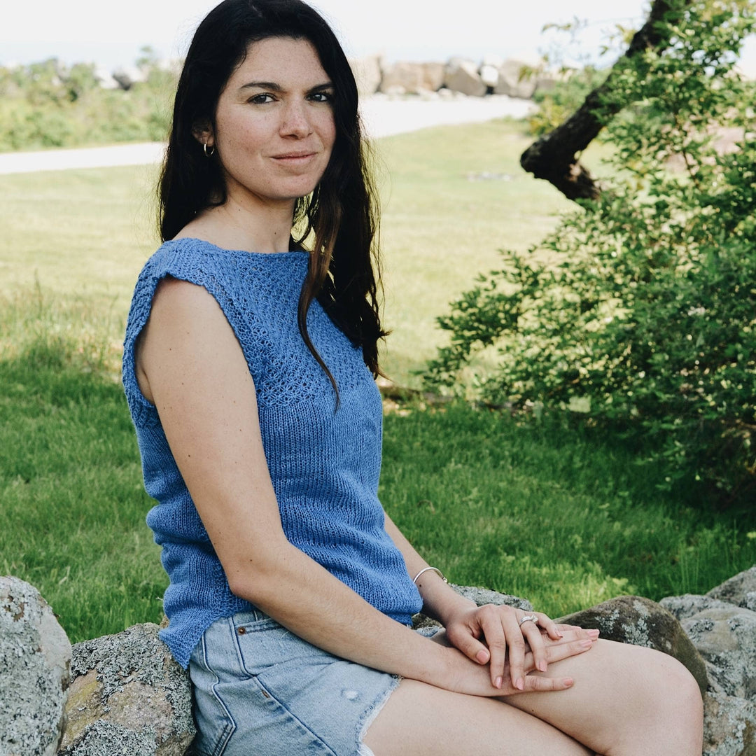 model wearing blue linen daisy stitch top while sitting on a rock wall with greenery in the background.