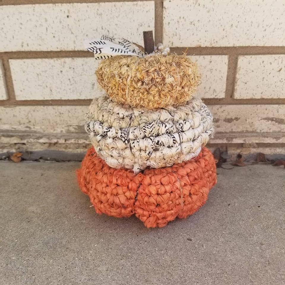 Stack of 3 crocheted pumpkins in yellow, beige and orange sitting against a brick wall