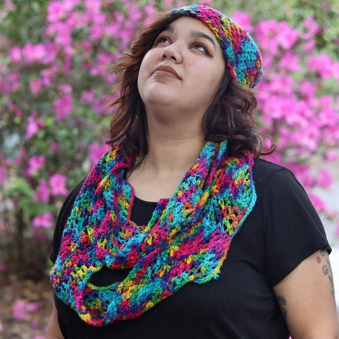 Close up of woman wearing a multicolored scarf and beanie set and a black t shirt standing in front of a pink floral bush
