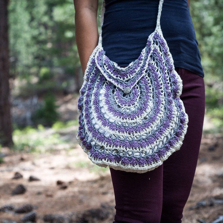 Purple and white mandala pattern crochet bag on a woman in navy tee shirt and purple pants standing in a wooded area