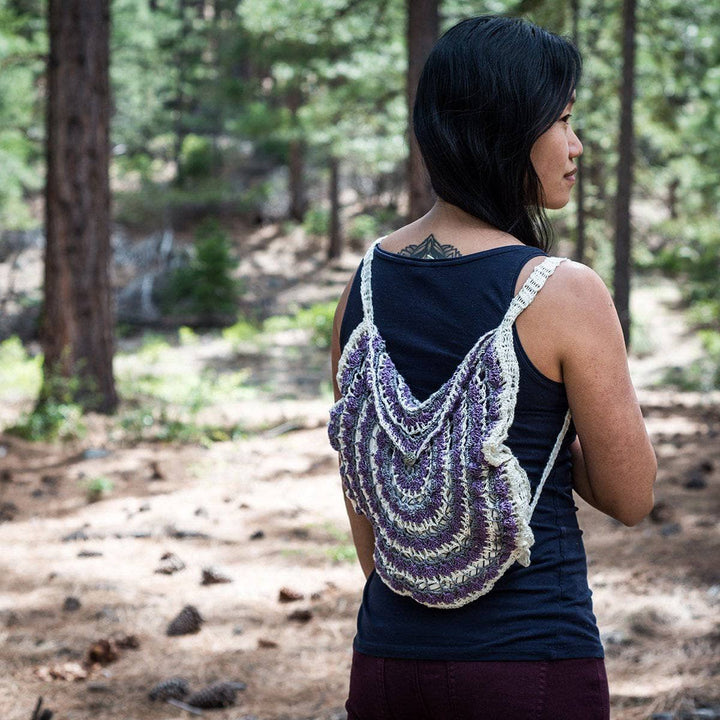 Back view of a purple and white mandala pattern crochet bag on a woman in navy tee shirt and purple pants standing in a wooded area