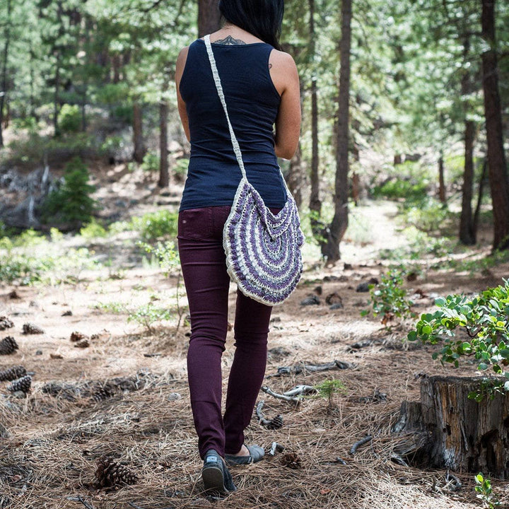 Purple and white mandala pattern crochet bag on a woman in navy tee shirt and purple pants walking in a wooded area