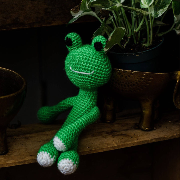 Completed Amigurumi Frog sitton on a wood shelf leaning against a brass planter and potted plant.
