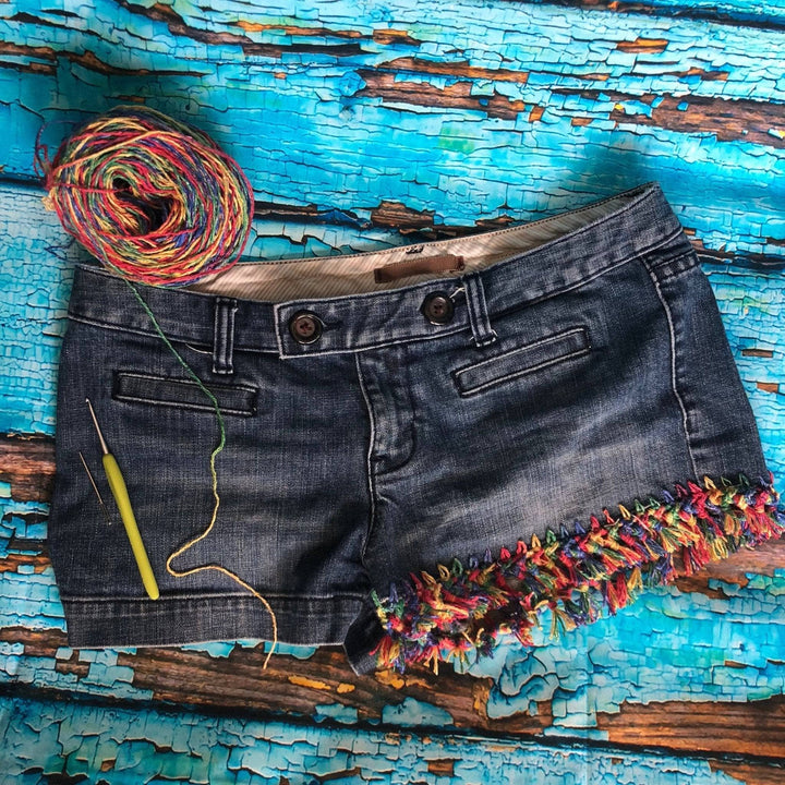 Jean shorts with DIY rainbow fringe, also visible is a skein of yarn, crochet hook, and needle. All on a wood background with flaking light blue paint.