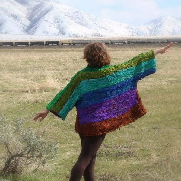 Back view of a woman wearing an oversized wide sleeve multicolor knitted jacket and standing in a grassy field