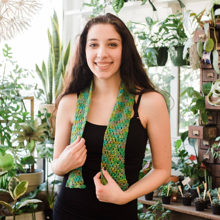 Model wearing crochet version of early spring scarf in crocodile snap with potted greenery in the background. 