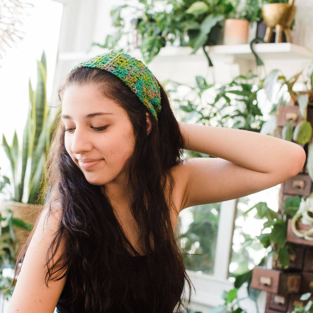 Model wearing crochet version of early spring scarf in crocodile snap as headband with potted greenery in the background. 