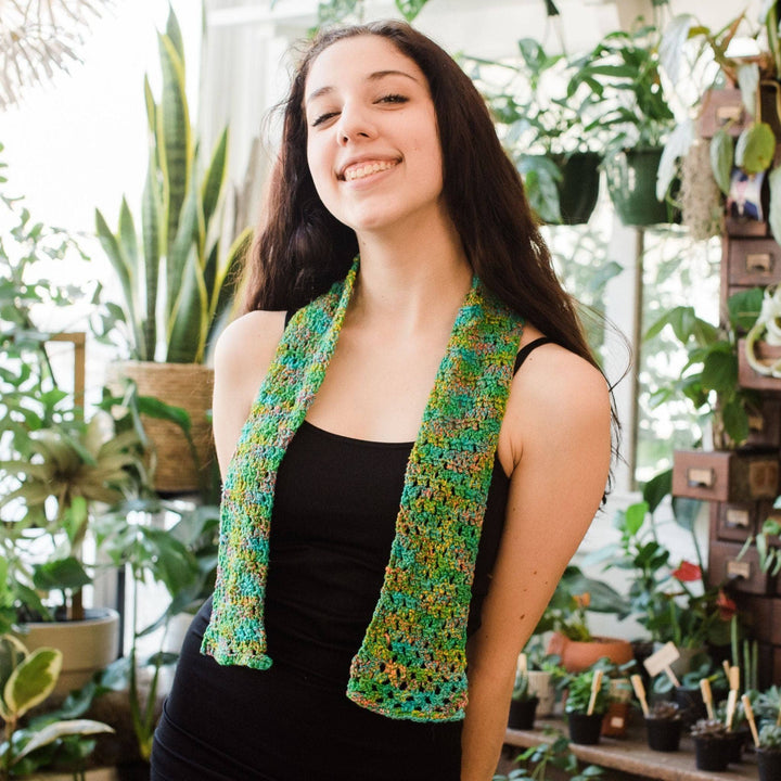 Model wearing crochet version of early spring scarf around neck with potted greenery in the background.