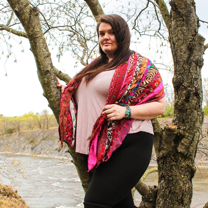 Model wearing earth festival scarf in pinks and reds on outside in front of a river. 