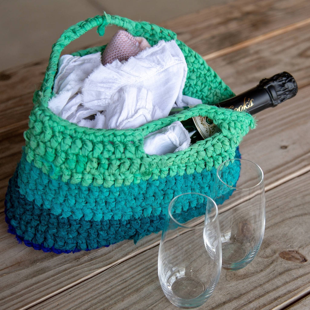 Crochet version of recycled chiffon market tote in ocean blues filled with napkins and wine bottle and wood background (wine not included)