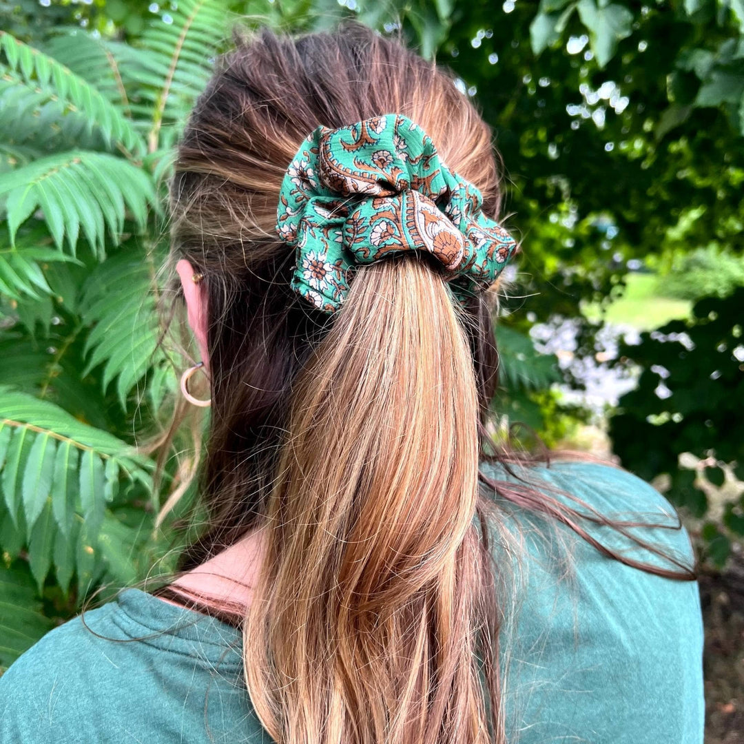 Model is wearing a green sari silk scrunchie in their pony tail  while standing outside in front of greenery 
