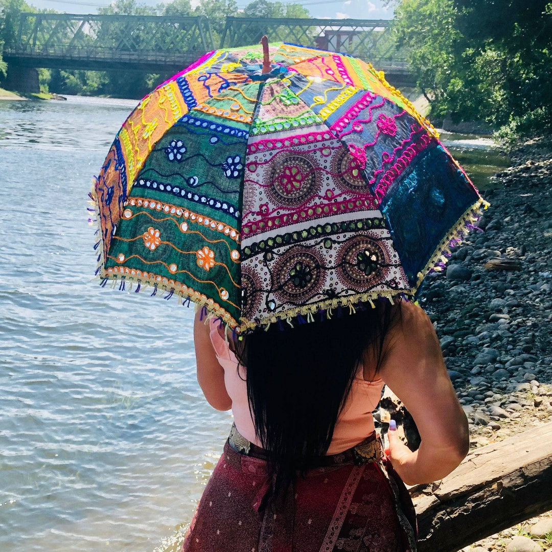 A woman standing by the water near a beach wearing a skirt made from recycled sari material holding an embellished parasol umbrella over her head.