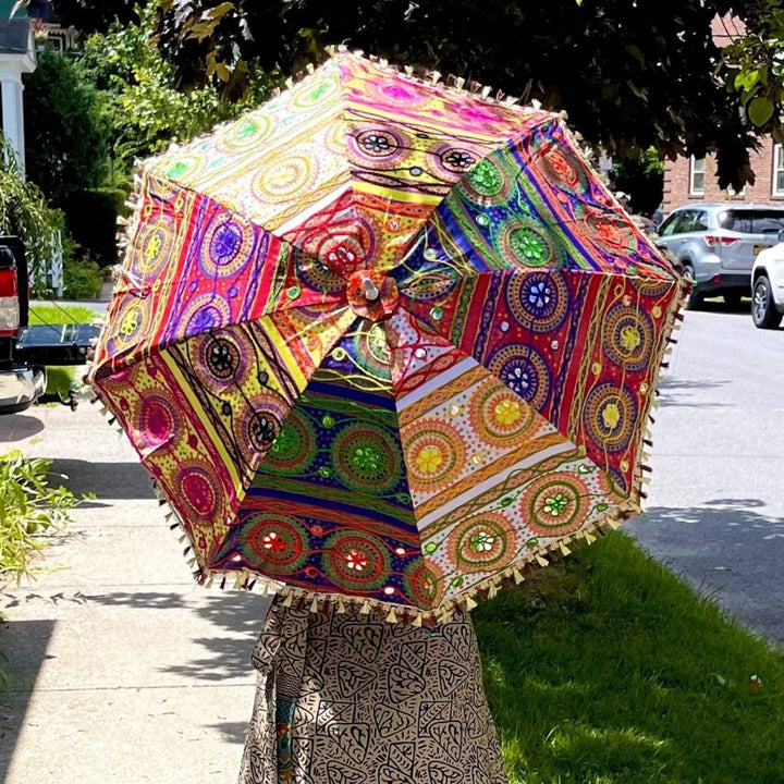 A woman walking down the street holding an embellished parasol. There's sun patterns all over it. It's bright and colorful with sparkles and shiny mirrors on it.