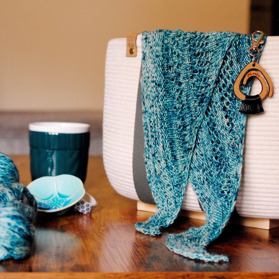 Emerald bay shawl hanging on the edge of a white basket with balls of yarn, mug, and small dish in the background.