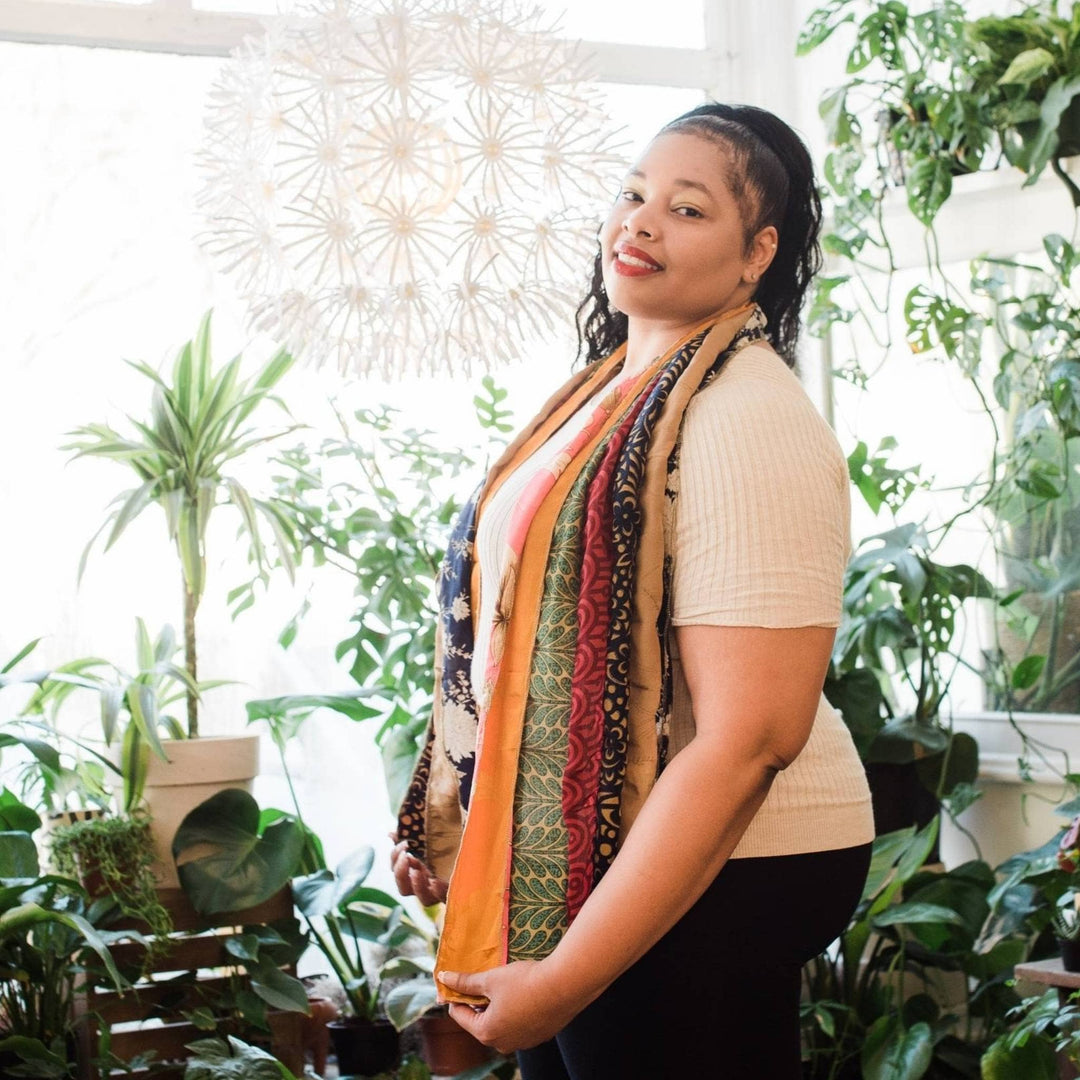 Model is standing in front of potted plants wearing an earthy toned colored medley scarf. 