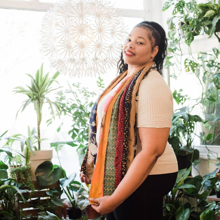 Model is standing in front of potted plants wearing an earthy toned colored medley scarf. 
