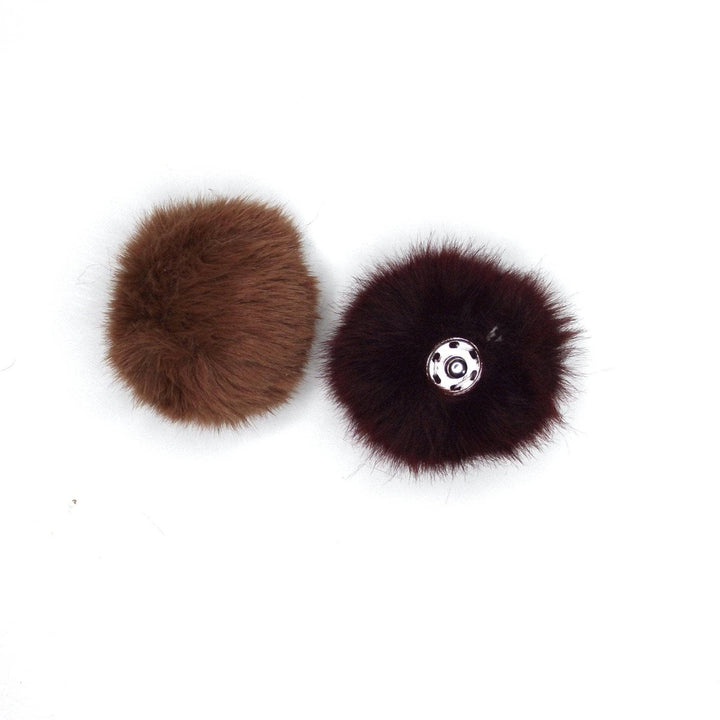 One light brown completed pom pom next to one upside-down completed dark brown pom pom with snap showing in front of a white background. 