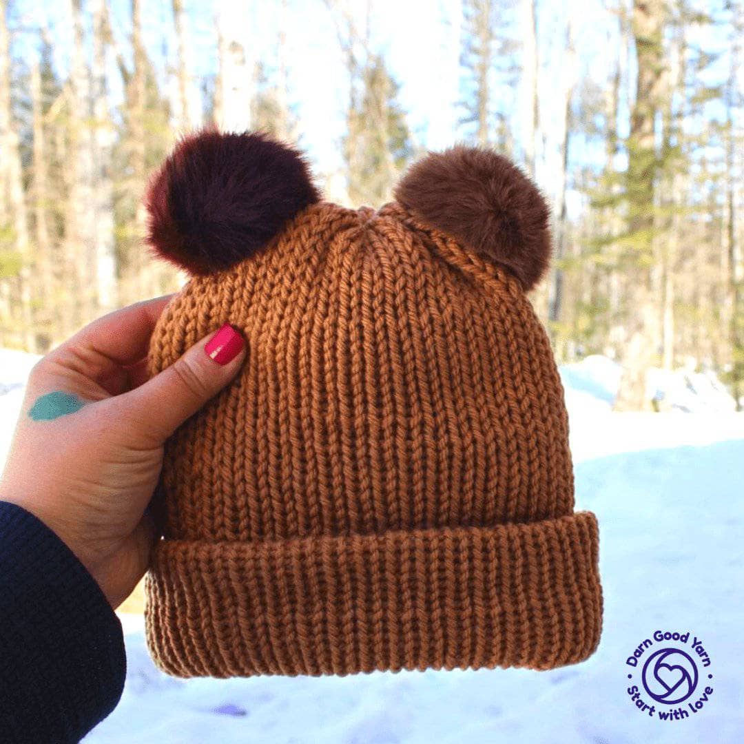 Brown knit hat with two faux fur pom poms being held by a hand in front of a winter woods background. 