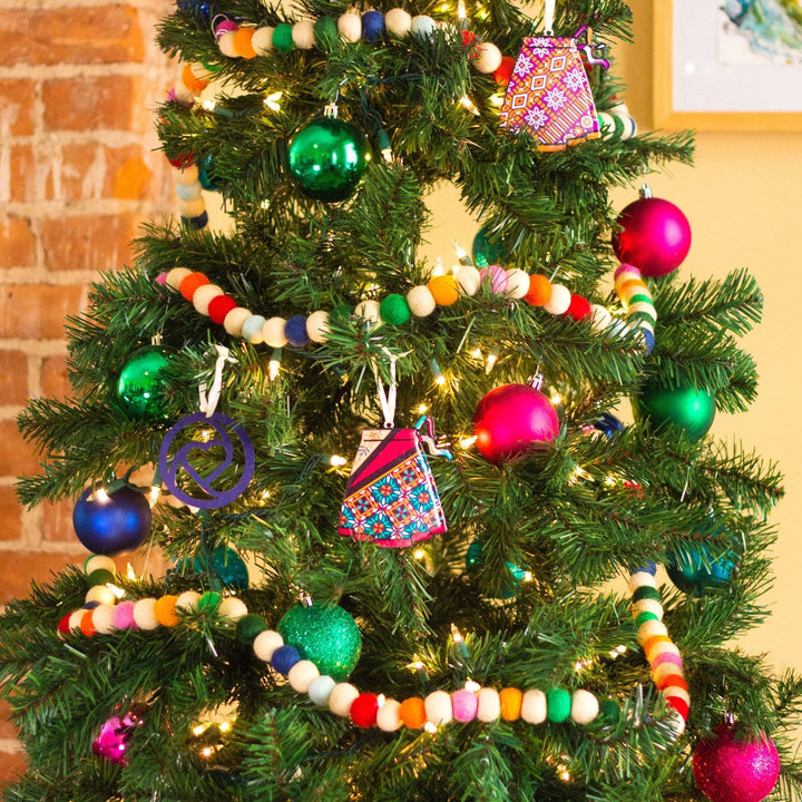 An up close photo of a christmas tree with rainbow felt ball garland and multi colored ornaments on it. There's also ornaments that look like sari wrap skirts.