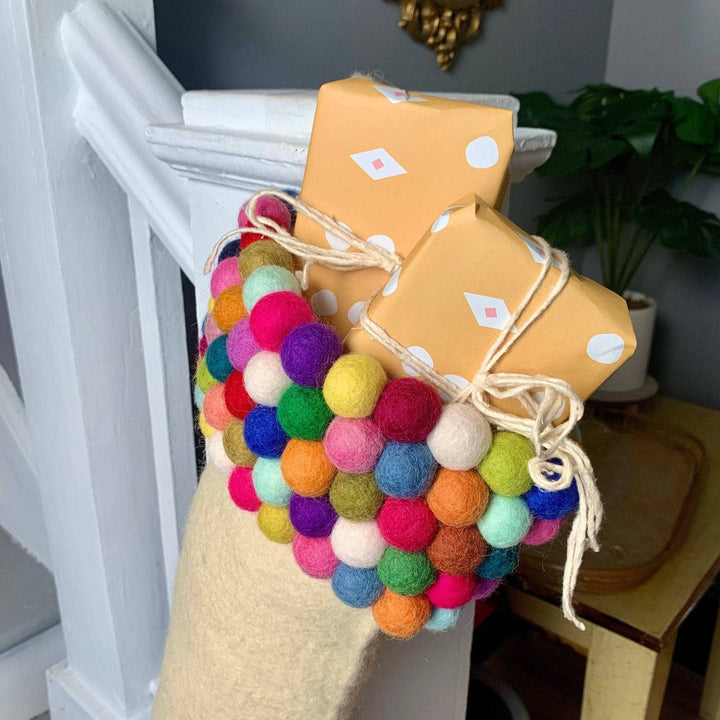 Close up imagery of hand made felt ball stocking. This image is a close up shot of the felt ball detailing at the top of the stocking and the contents in the stocking.Light cream colored felt stocking. The top of the stocking consists of  five rows of colorful felt balls. The stocking is holding two small boxes wrapped in yellow paper with white detailing and tied in a bow with off white yarn. The stocking is hung on a white railing of a set of stairs.