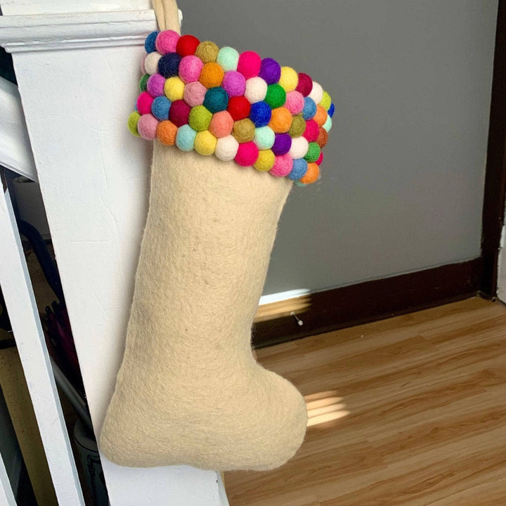 Light cream colored felt stocking. The top of the stocking consists of five rows of colorful felt balls.  The stocking is hung on a white railing of a set of stairs.