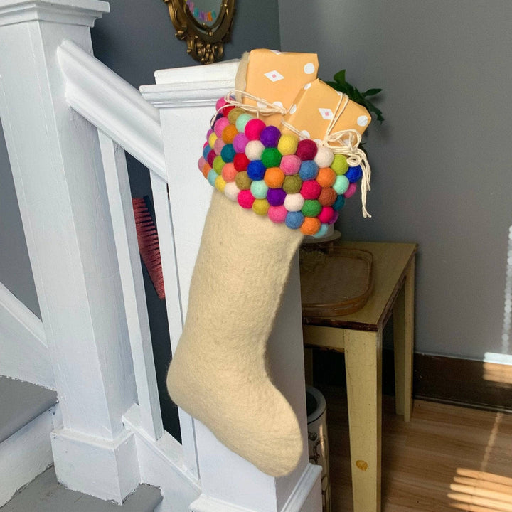 Light cream colored felt stocking. The top of the stocking consists of  five rows of colorful felt balls. The stocking is holding two small boxes wrapped in yellow paper with white detailing and tied in a bow with off white yarn. The stocking is hung on a white railing of a set of stairs.