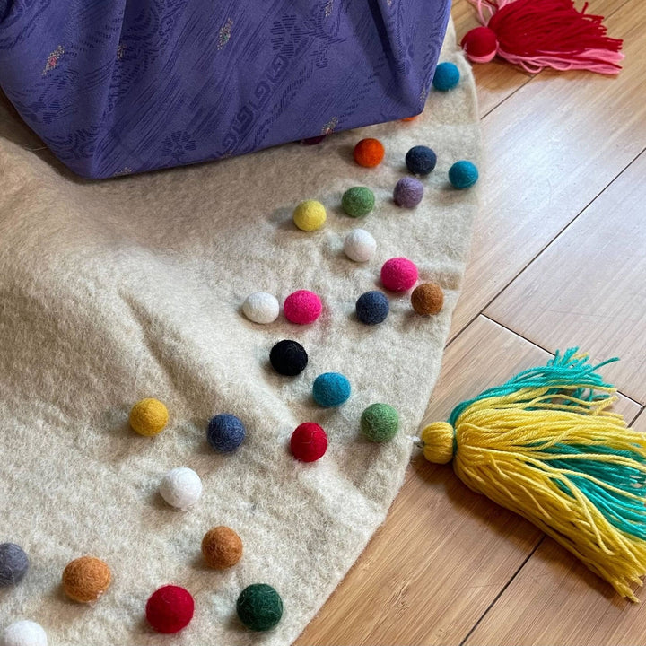 An up-close view of the felt ball tree skirt. There's rainbow felt balls on the top of the cream colored skirt with various colored tassels on the edges of the skirt.