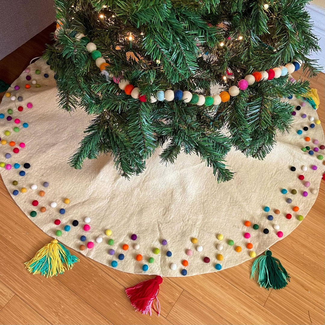 An above view of a cream colored felt ball christmas tree skirt. There's felt ball garland on the christmas tree above. The felt balls on the skirt are rainbow colored and are assorted along the edges