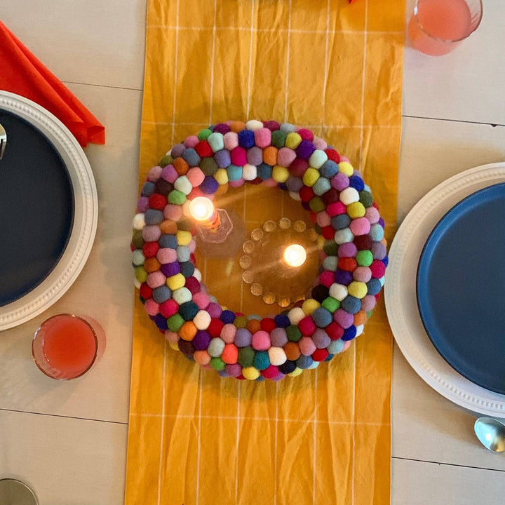 Overhead image of a multicolored circular wreath made out of felt balls set on top of a white table with yellow striped runner. The wreath is being used as a center piece featuring 2 colorful patterned tapered candles in glass candle holders. The candles are lit and the place setting is paired with two navy plates and gold cutlery.