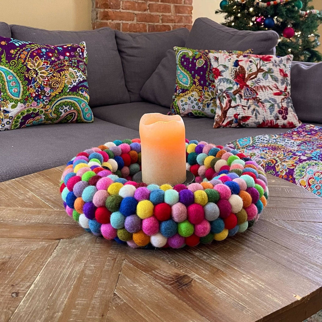 A Felt Ball wreath used as a centerpiece on a coffee table. There's a lit pillar candle in the center and kantha quilt pillows on the couch behind it.