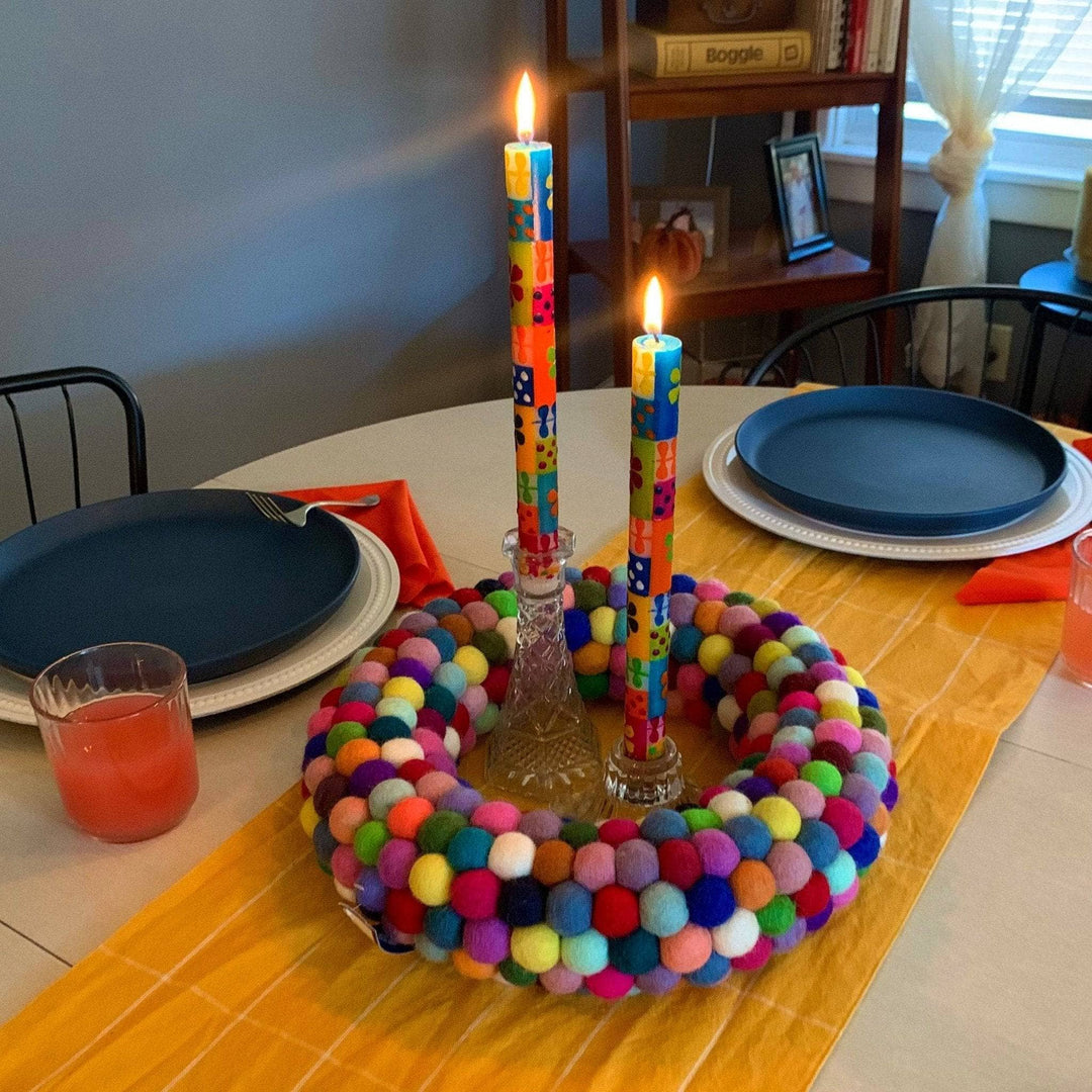 Multicolored circular wreath made out of felt balls set on top of a white table with yellow striped runner. The wreath is being used as a center piece featuring 2 colorful patterned tapered candles in glass candle holders. The candles are lit and the place setting is paired with two navy plates and gold cutlery. 