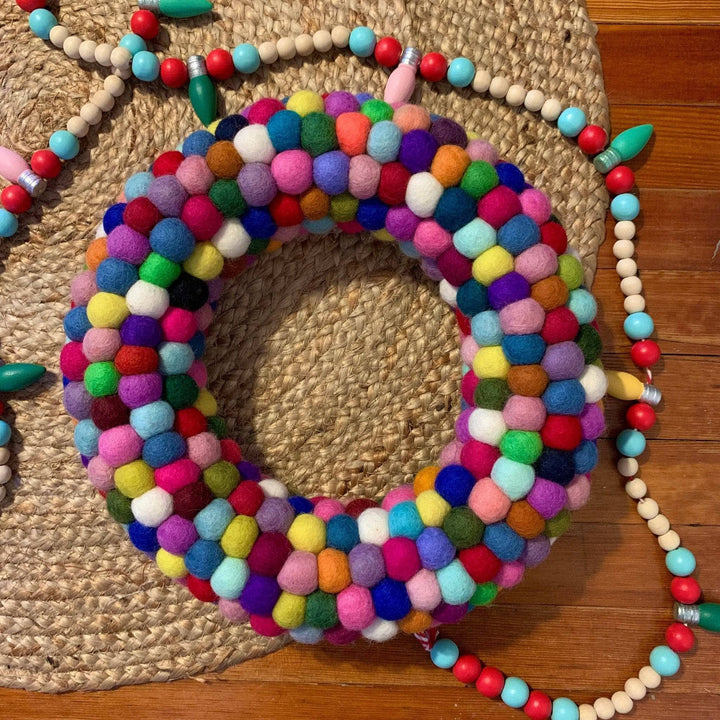 Multicolored circular wreath made out of felt balls laid on top of a woven rattan like material and a wooden slated board. The wreath is surrounded by some extra festiveness in the form of a wooden beaded garland featuring red, blue, green yellow and natural wooden beads and lightbulbs.