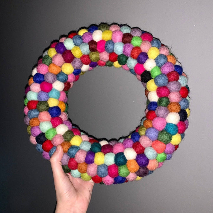 Multicolored circular wreath made out of felt balls being held against a gray wall