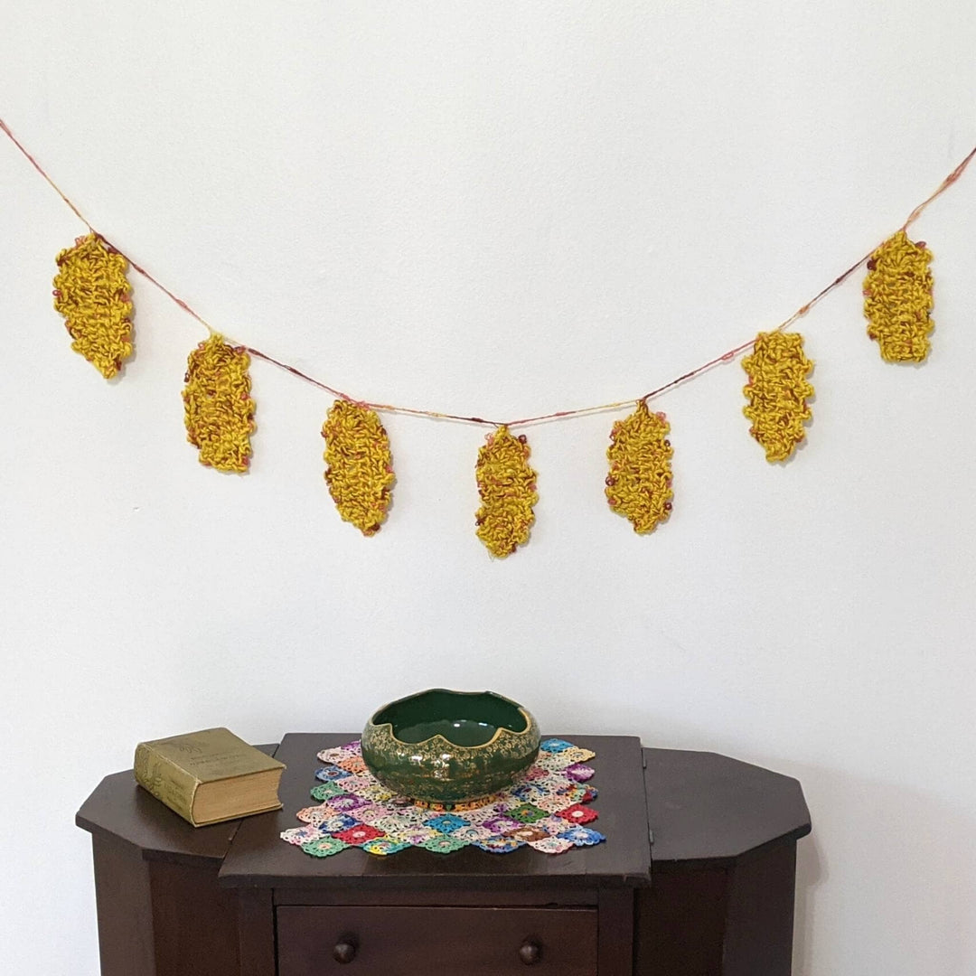 Festive fall garland hung up on a white wall with dark wood table holding doily, book, and bowl below.