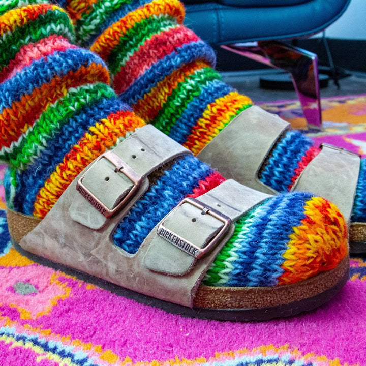 Model is wearing colorful fleece lined wool socks with sandals while standing on a pink carpet.  