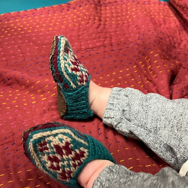 Model wearing Folklore Handmade slippers in baby size sitting on a red blanket.
