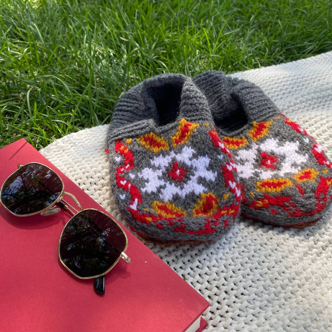 Grey, red and yellow folklore slippers are sitting on a blanket in the grass next to a book and sunglasses. 