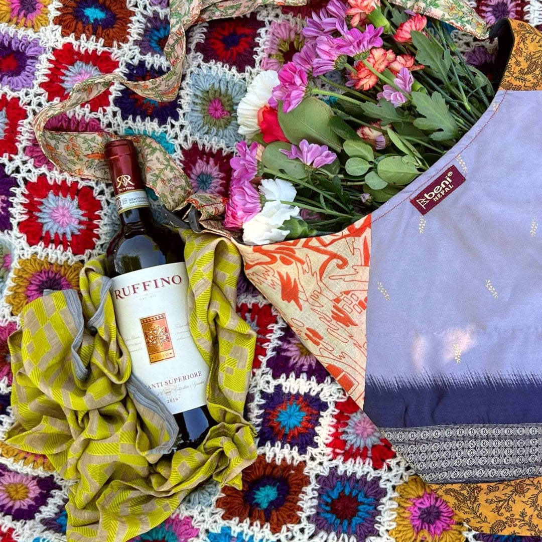 Green furoshiki is laying under a wine bottle on a crochet blanket in the park next to a sari silk purse with flowers in it. 