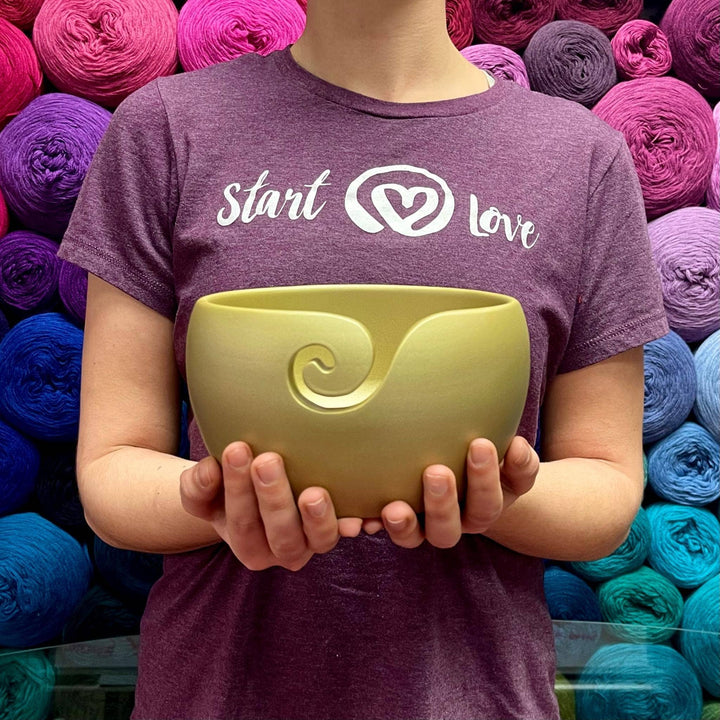 Model holding gold finished wooden yarn bowl while wearing purple start with love t-shirt and multicolor yarn background.