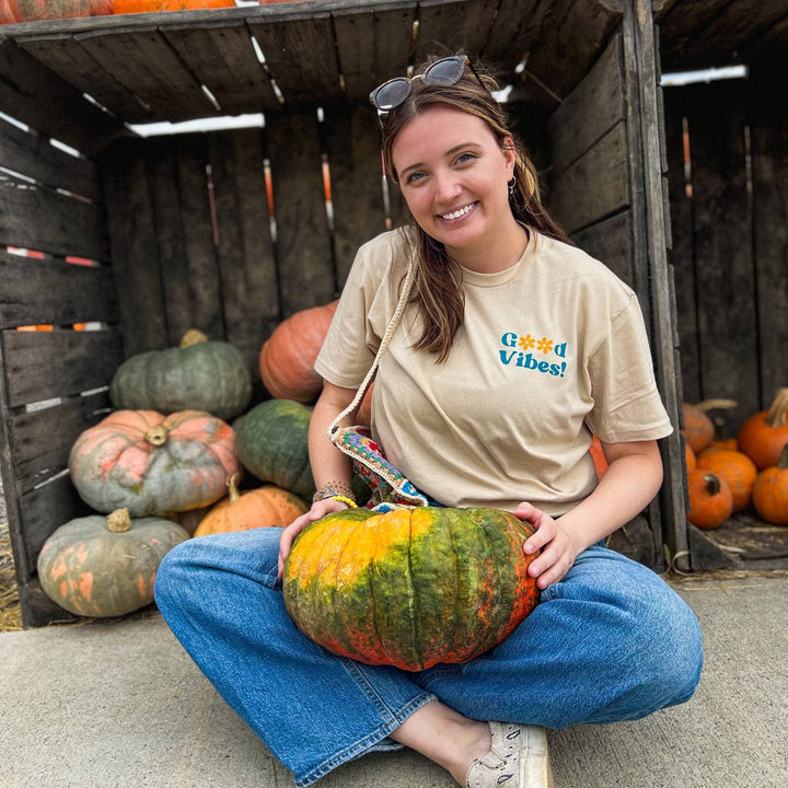 Model is sitting in front of a crate of pumpkins wearing the Good vibes shirt . 