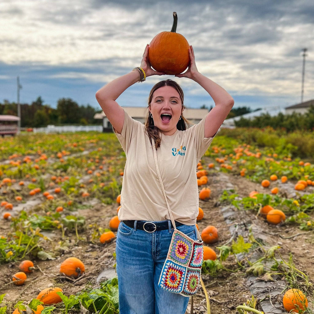Model is holding a pumpkin above her head wearing a good vibes shirt and granny square bag over her shoulder. 
