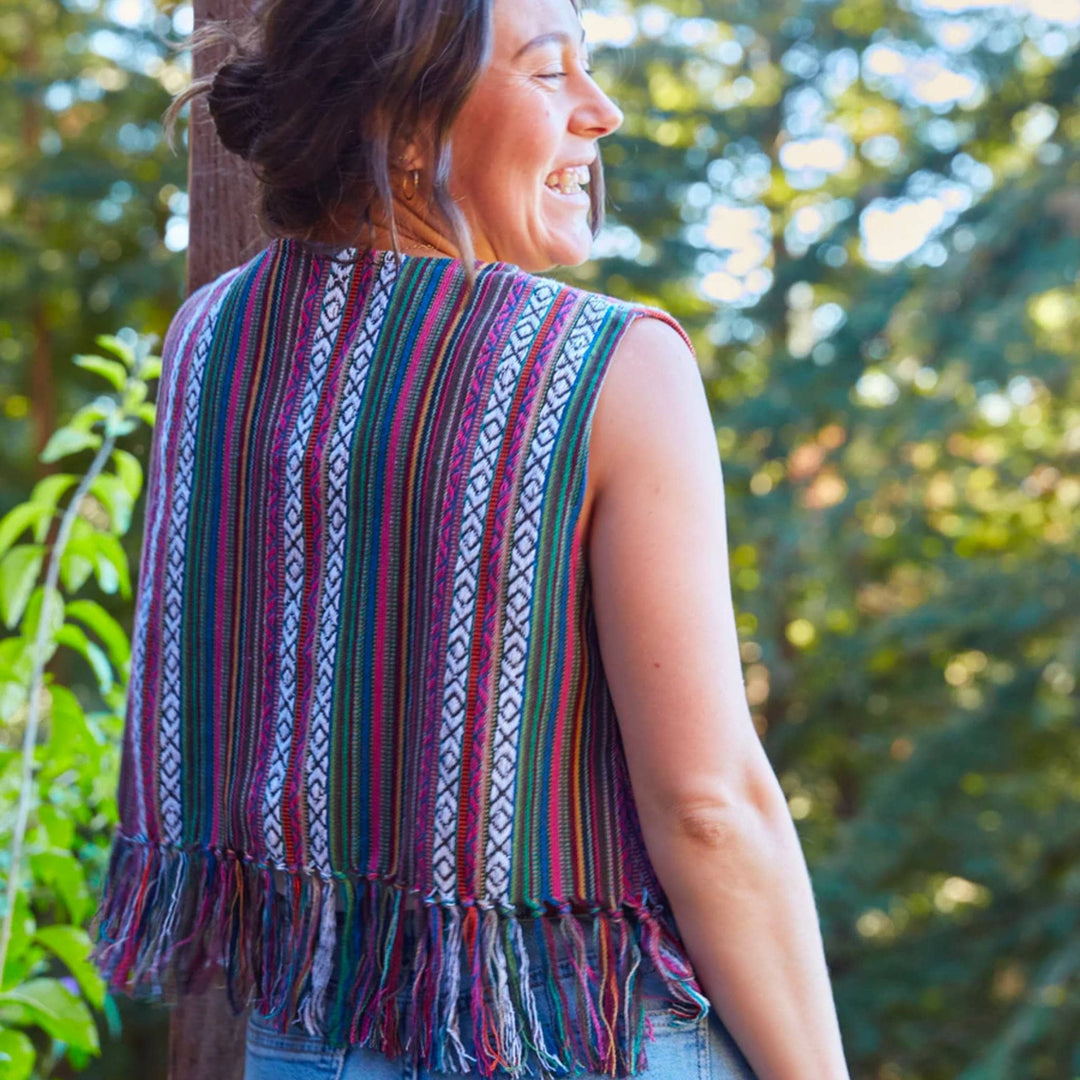Model is outside in front of some trees with their back facing the camera wearing a lavender heather bolero vest. 