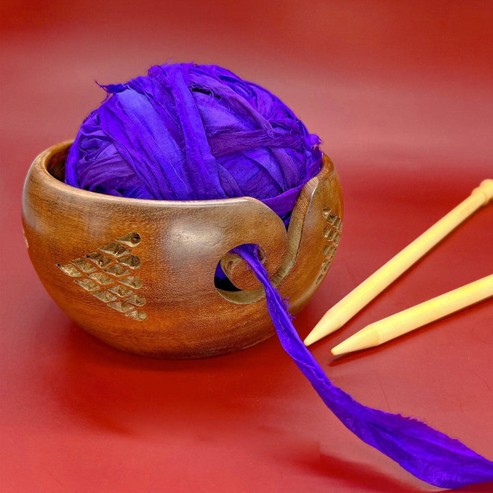 Geometric wooden yarn bowl with skein of sari silk ribbon yarn ultra violet in it and a set of knitting needles beside.