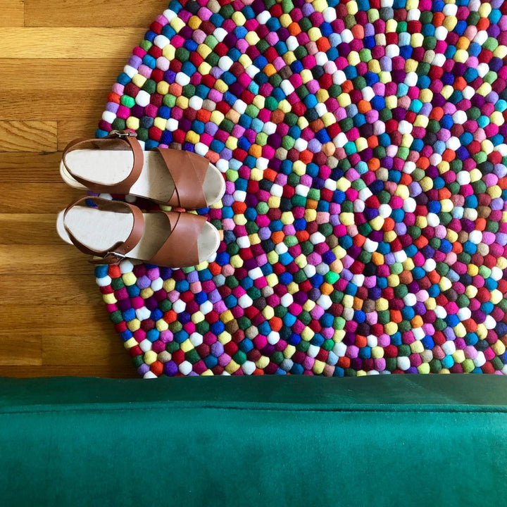 Handcrafted Limited Edition Felt Ball Rug on a wood floor under a green couch with brown sandals laying on them. 