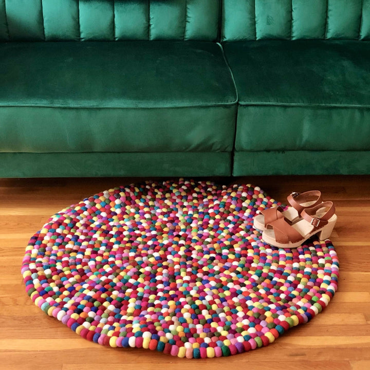 Multicolored handcrafted Limited Edition Felt Ball Rug on a wood floor under a green couch with brown sandals laying on them.