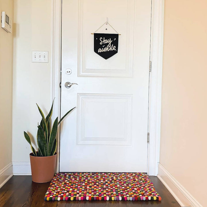 Small rectangle felt ball rug in front of a door with a plant next to it. 