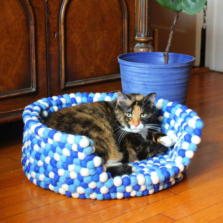 A Calico cat sitting in a blue & white felt ball pet bed. The balls are white, dark blue and light blue.