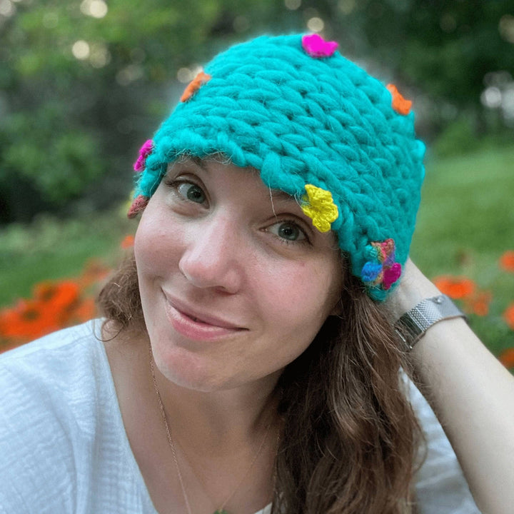 Founder Nicole wearing flower crown wool headband  outside with blurred greenery in the background. Headband is a wide strip of knitting with small brightly colored flowers throughout.