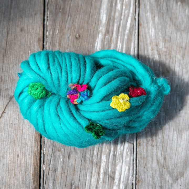 Single skein of blue/green wool yarn with multicolor handmade silk flowers attached sitting on a wood background. 